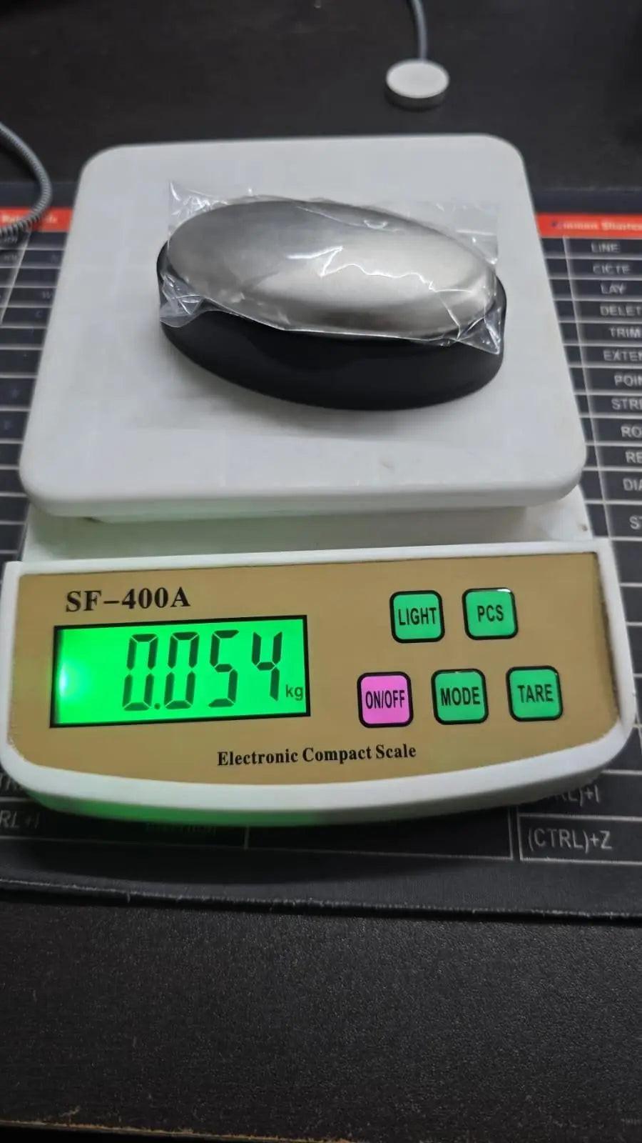 The Odor Remover Hand Bar Stainless Steel Soap, a small silver oval wrapped in plastic, sits on an SF-400A Electronic Compact Scale showing a weight of 0.054 kg (54 grams).