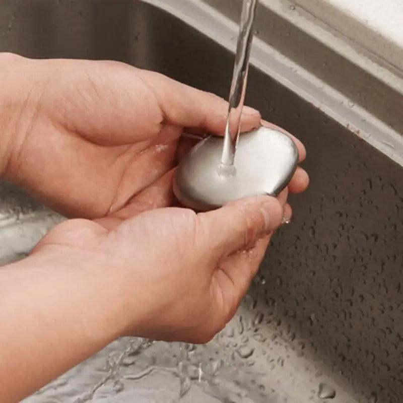 Someone uses the Odor Remover Hand Bar Stainless Steel Soap under running water at the sink, washing both hands to eliminate unwanted odors.